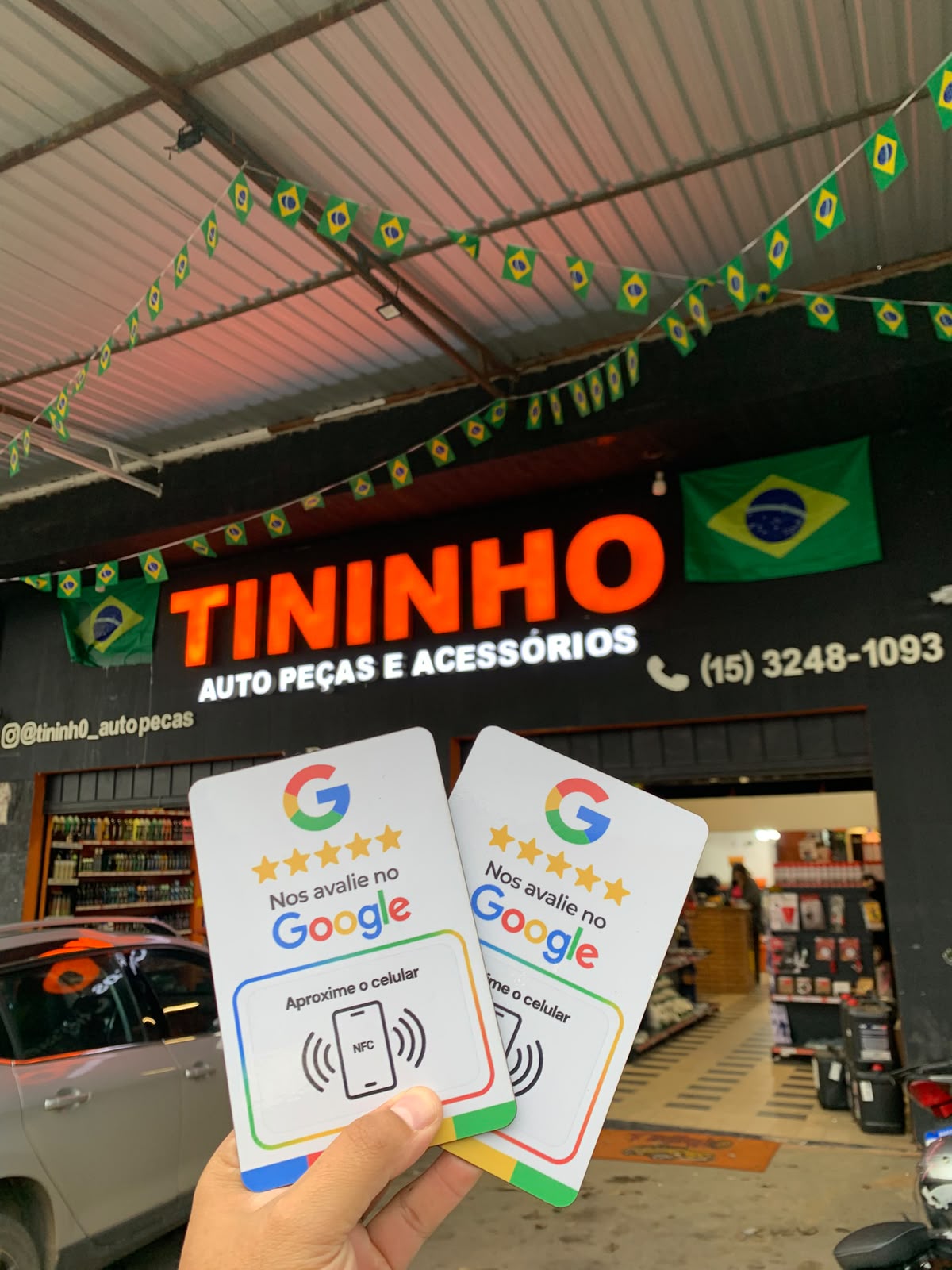 Mão segurando duas placas de avaliação em frente à loja Tininho Auto Peças