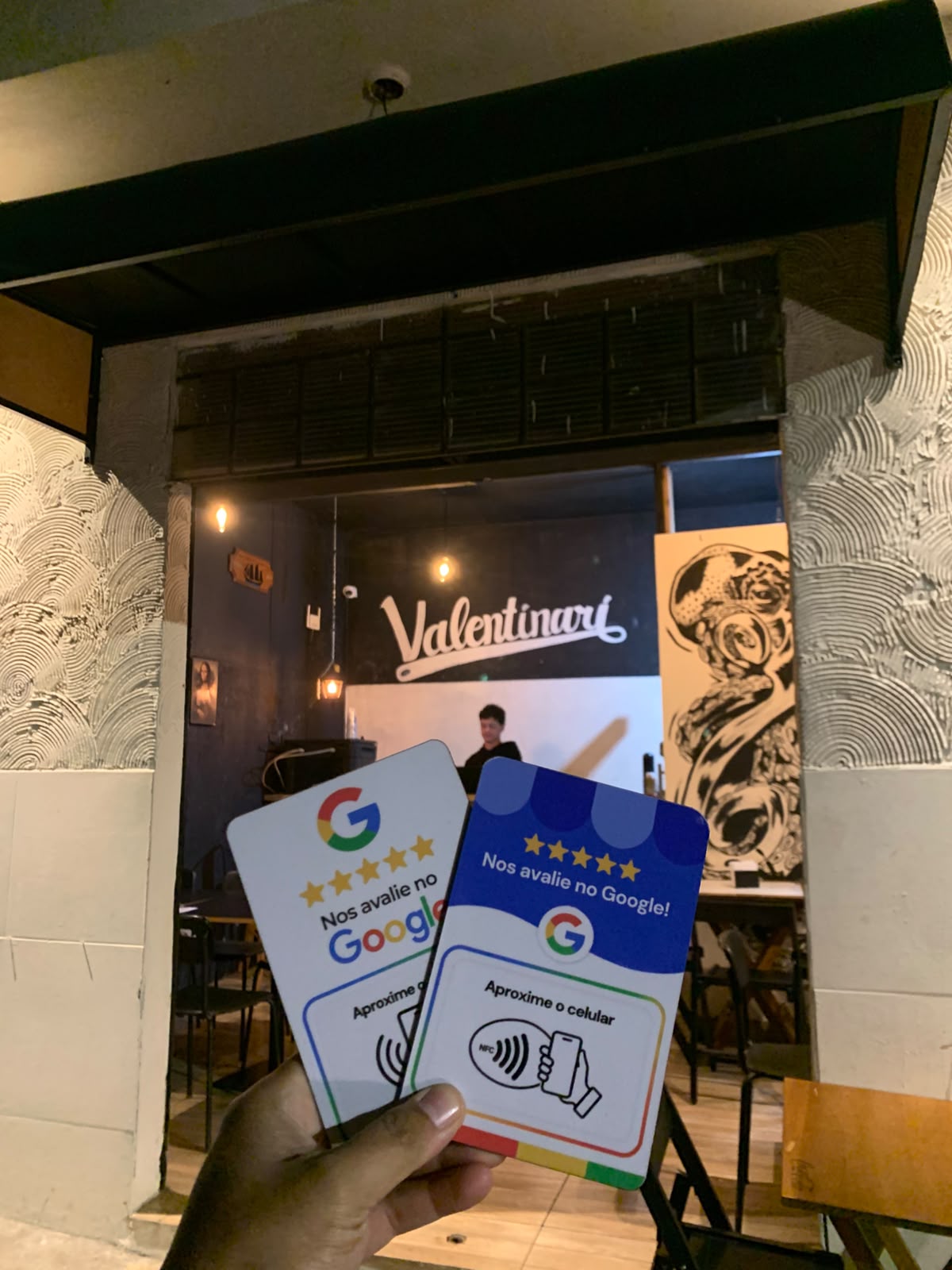 Mão segurando duas placas de avaliação em frente à fachada do Valentinari à noite